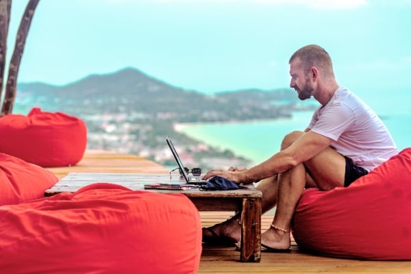 A man sitting on a red cushion working remotely with his laptop. There are mountains and the sea in the background